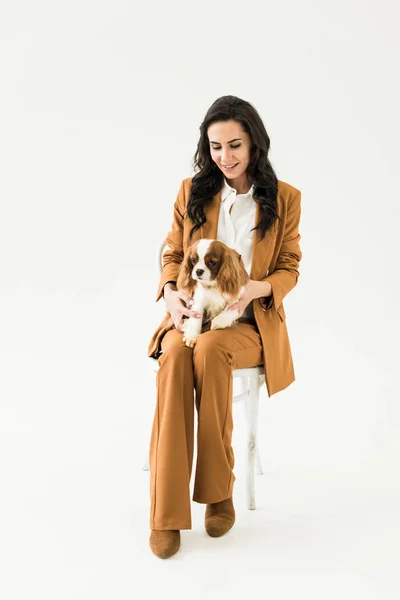 Smiling young woman in brown suit sitting on chair and holding dog on white background — Stock Image Smiling Young Woman Brown Suit Sitting Chair Holding Dog White Stock Photo