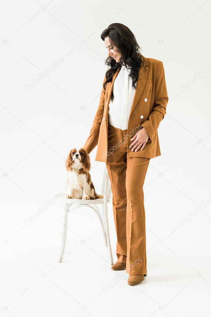 Pregnant woman in elegant brown suit stroking dog on white background