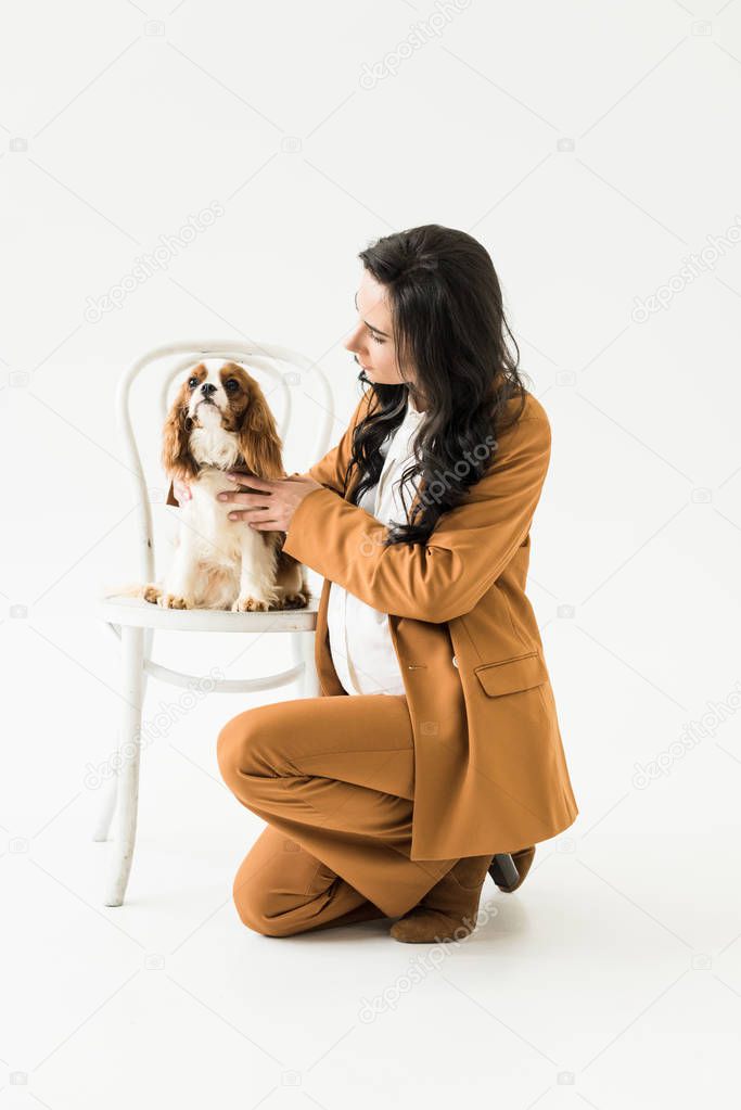 Pregnant woman sitting near chair and stroking dog on white background