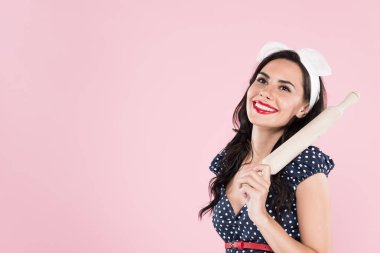 Laughing brunette woman in dotted dress holding wooden rolling pin isolated on pink clipart