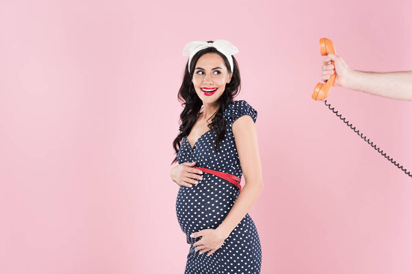 Смеющаяся беременная женщина смотрит на телефонный приемник, изолированный на розовый

