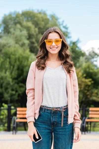 pretty girl in sunglasses smiling while holding smartphone in park