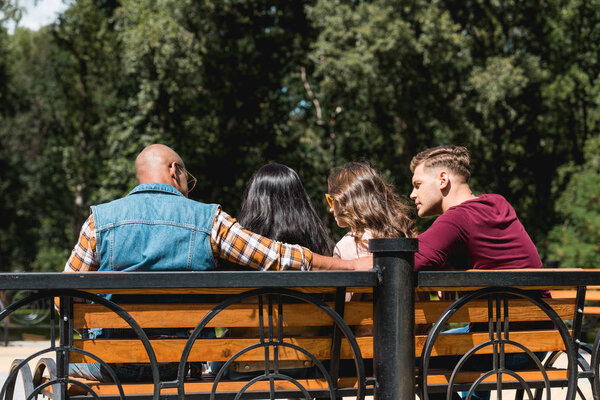 back view of multiethnic friends sitting on bench in park