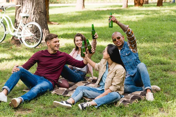 happy multiethnic group of friends sitting on plaid blanket and clinking bottles in park