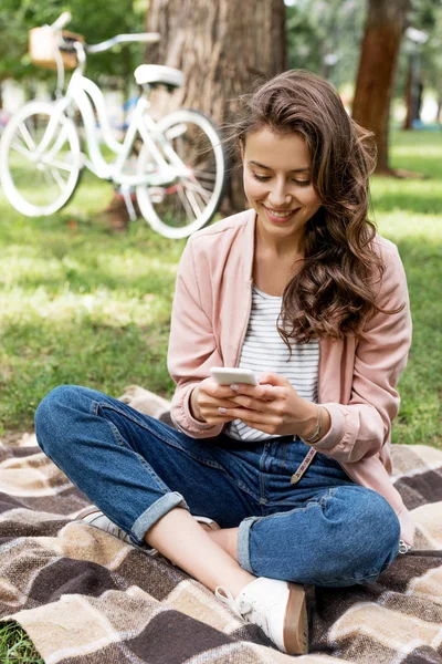 欢快的女孩看着智能手机 而坐在格子毯子在公园 — 图库照片