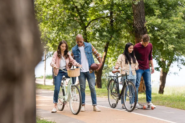 可愛い女の子が乗馬 自転車や公園内の陽気な友人ロングボーディング近くに歩いてアフリカ系アメリカ人の男 — ストック写真
