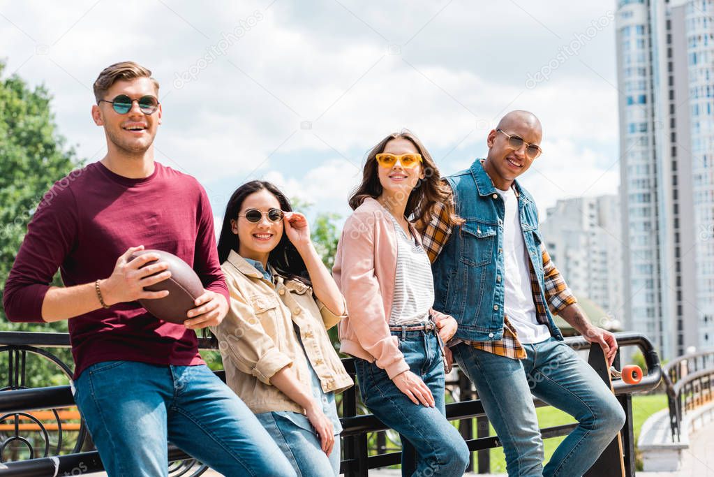 happy multicultural young men in sunglasses standing with longboard and american football near attractive girls