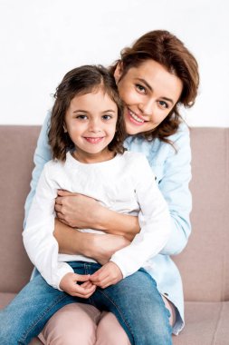 Laughing mother sitting on sofa and holding daughter on knees isolated on white  clipart
