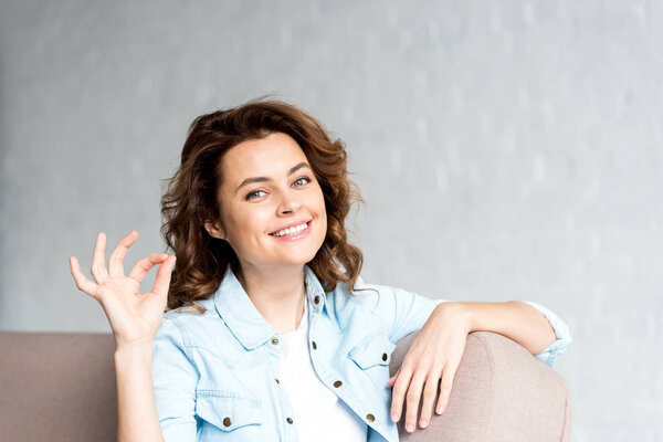 Happy curly woman in shirt sitting on sofa and showing okay sign on grey 
