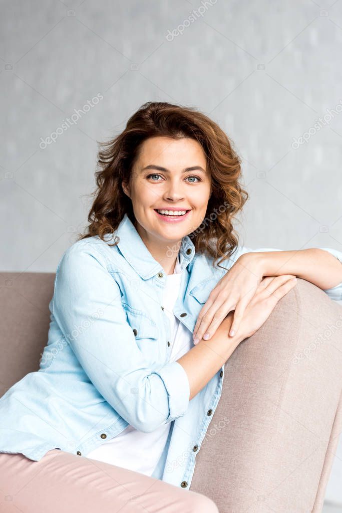 Cheerful curly woman in shirt sitting on sofa and smiling on grey 