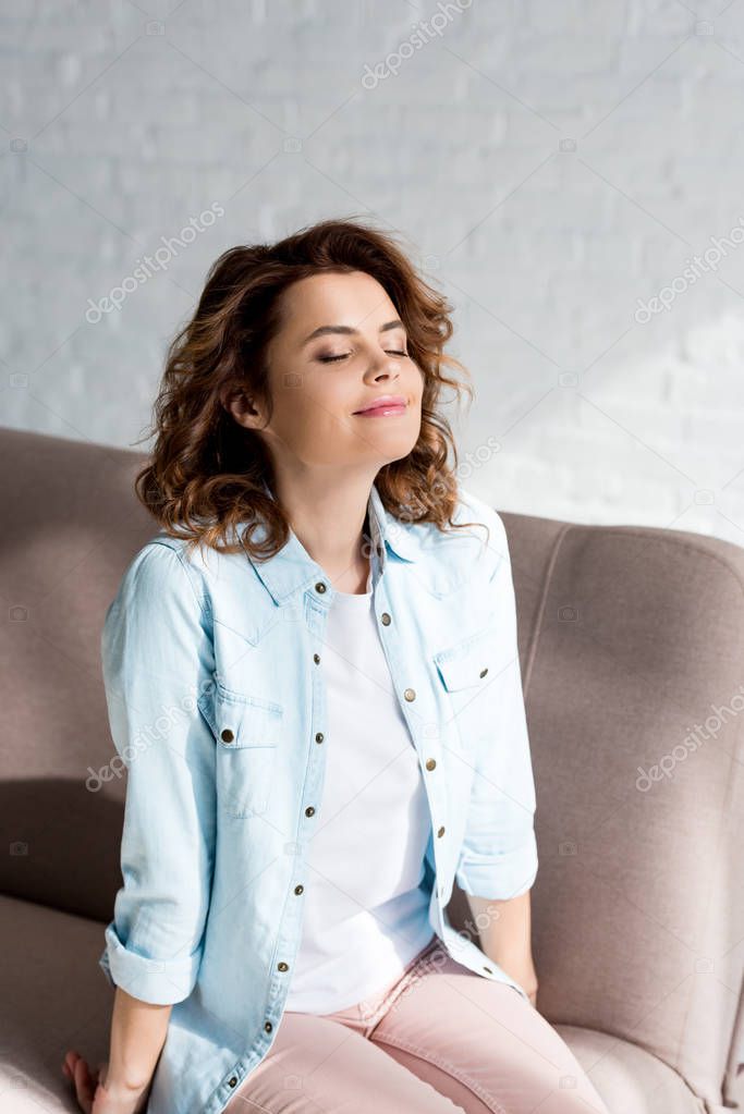 Inspired curly woman in shirt sitting on sofa with closed eyes on grey 