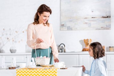 Smiling woman with wicket basket looking at daughter while serving table for easter clipart