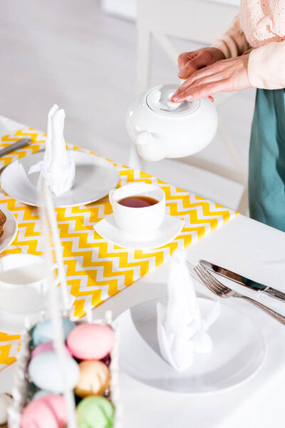 Partial view of woman with teapot pouring tea in cup on table
