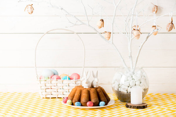 Пасхальный торт с керамическими кроликами и плетеная корзина с окрашенными яйцами на столе на деревянном
 