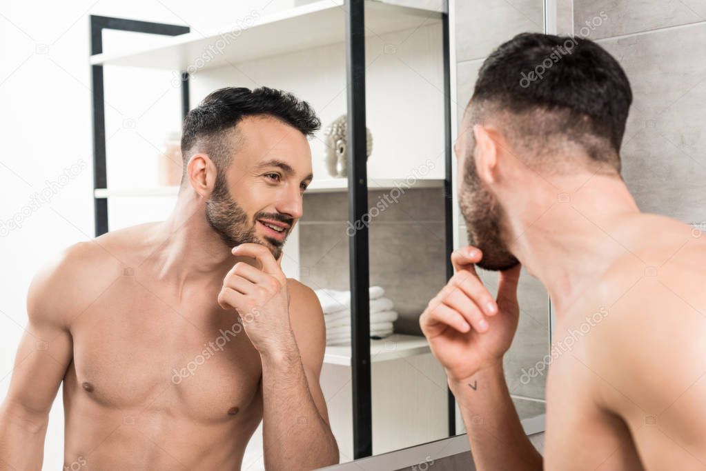 handsome shirtless man looking at mirror and touching face in bathroom 