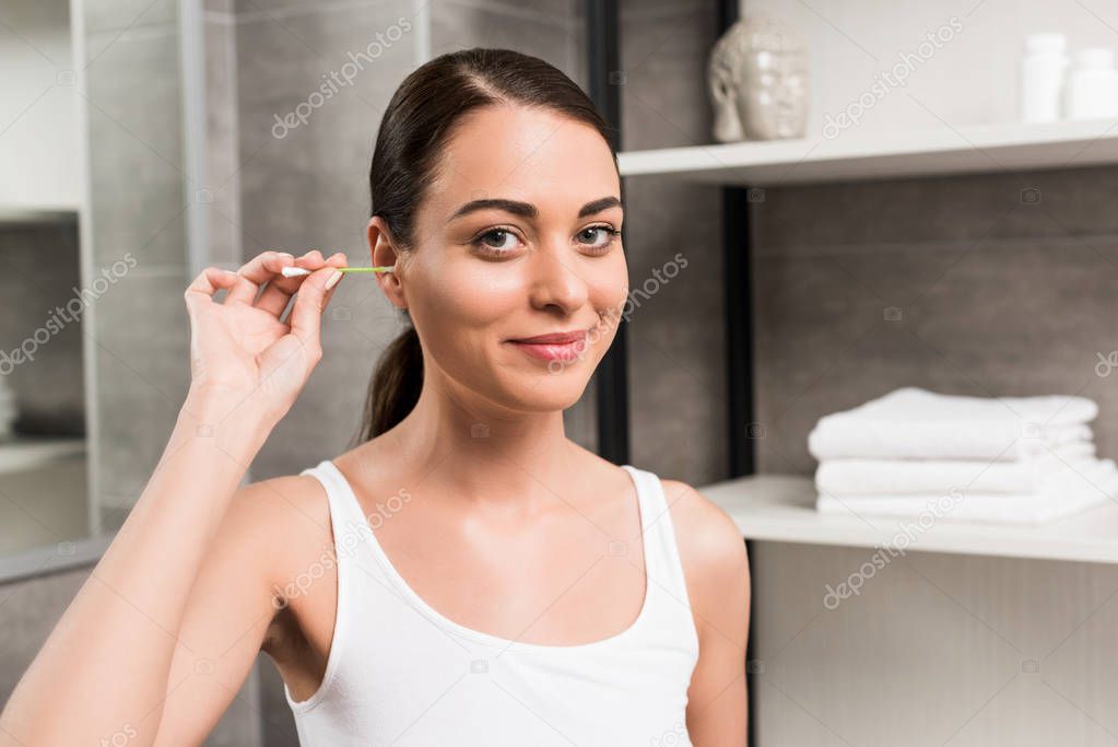 attractive brunette woman smiling while cleaning ear in bathroom 