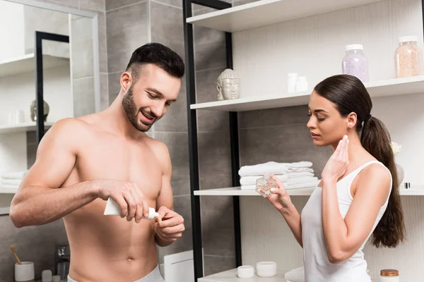 魅力的な女性の香水ボトルを押し 歯ブラシと歯磨き粉の浴室で上半身裸の彼氏の近くに立っている間に適用すること — ストック写真