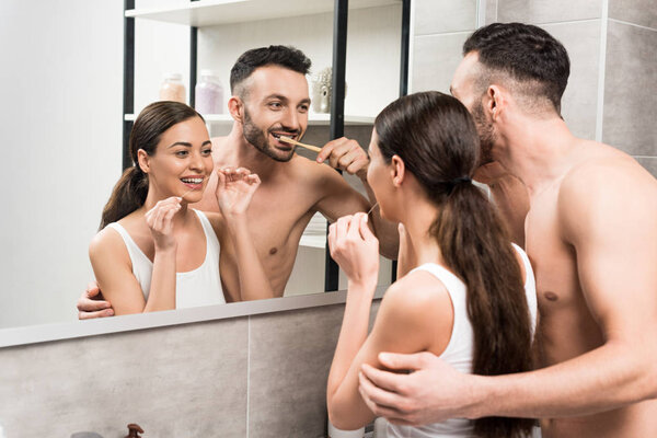bearded boyfriend brushing teeth near girlfriend using dental floss while looking at mirror in bathroom 
