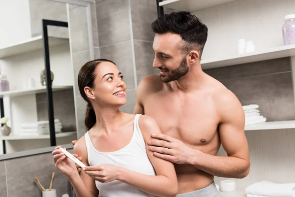 バスルームでの妊娠検査を保持している陽気なガール フレンドを見て満足している上半身裸の男 — ストック写真