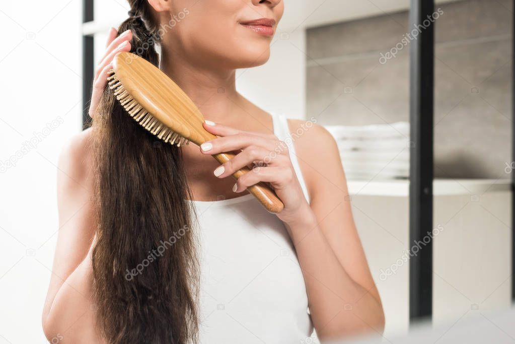 cropped view of happy brunette woman brushing hair while standing in bathroom 