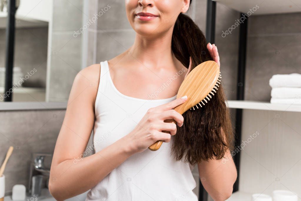 cropped view of smiling brunette woman brushing hair while standing in bathroom 