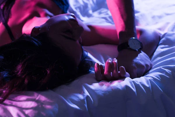 ネオンライトが付いているベッドで情熱的な女性の手を繋いでいるセクシーな男のビューをトリミング — ストック写真
