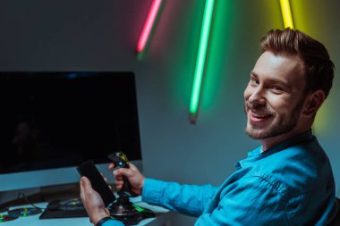 selective focus of smiling and handsome man holding joystick and smartphone  clipart