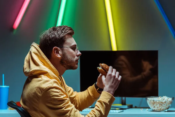 Vista Lateral Hombre Adulto Guapo Joven Comiendo Sabrosa Hamburguesa Mirándolo —  Fotos de Stock