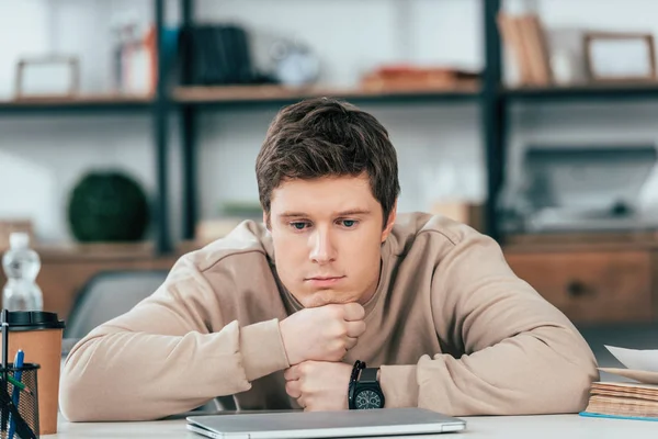 悲伤的学生在手表坐在折叠的手臂靠近笔记本电脑 — 图库照片