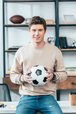 Masada oturan ve işyerinde futbol topu tutan gülümseyen genç adam