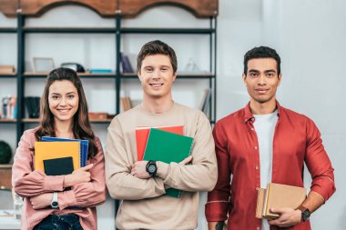 Defter ve kitap tutan ve kameraya bakan üç çok ırklı öğrenci