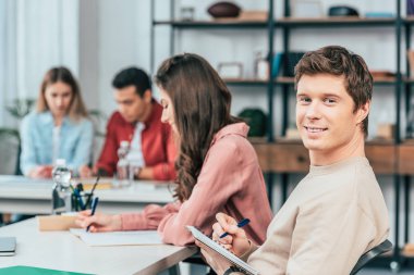 Mutlu öğrenci kalem ile defter tutarak ve gülümseme ile kamera bakıyor