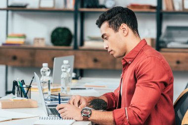 Concentrated student with tattoo in red shirt using laptop at desk clipart