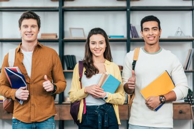 Notebooklar tutan ve başparmak yukarı gösteren üç çok etnik mutlu öğrenciler