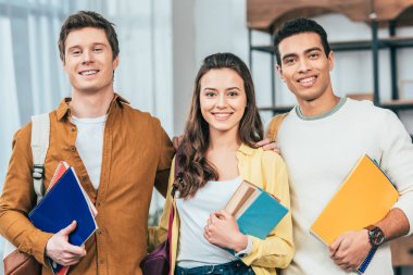 Defter ve kitap tutan ve kameraya bakan üç mutlu çok ırklı öğrenci