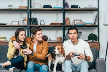 Three multicultural friends with beagle dog holding joysticks and playing video games clipart