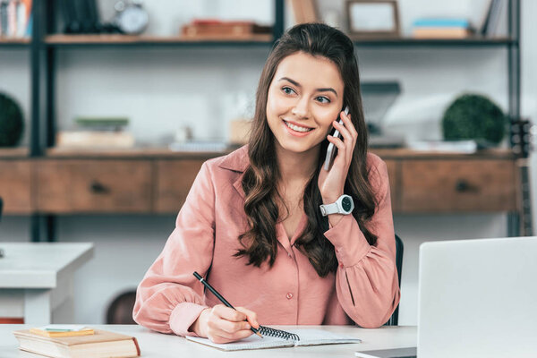 Темноволосая улыбающаяся девушка в розовой рубашке разговаривает на смартфоне и пишет в блокноте за столом с ноутбуком
