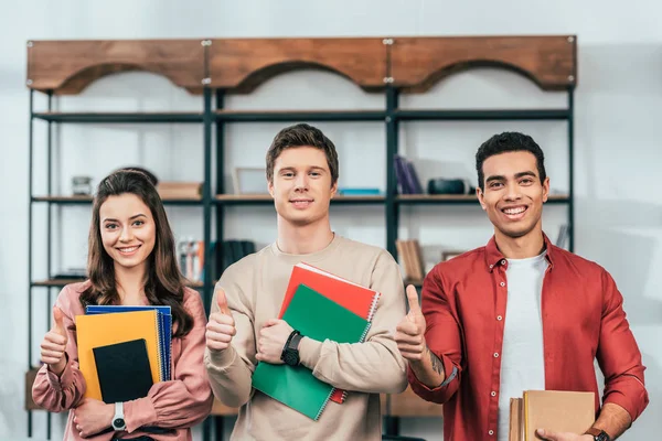 Tres Alegres Estudiantes Multiétnicos Sosteniendo Cuadernos Libros Mostrando Pulgares Hacia — Foto de Stock