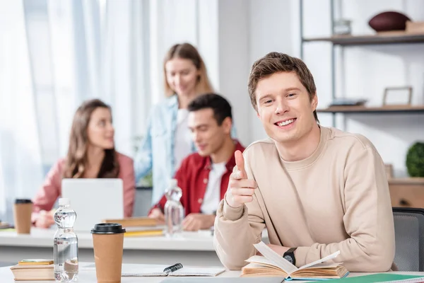 Estudiante Sonriente Con Libro Sentado Mesa Señalando Con Dedo Cámara — Foto de Stock