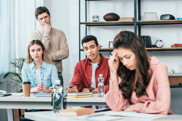 Worried multiethnic students sitting at table and looking at sad brunette girl