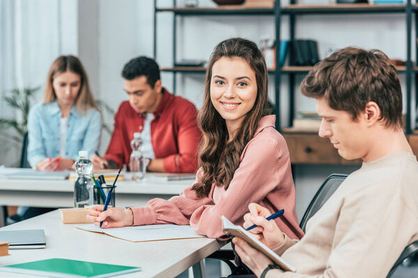 Charming student writing in notebook and looking at camera with smile