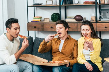 Kanepede oturan ve birlikte pizza yeme üç çok ırklı neşeli arkadaşlar