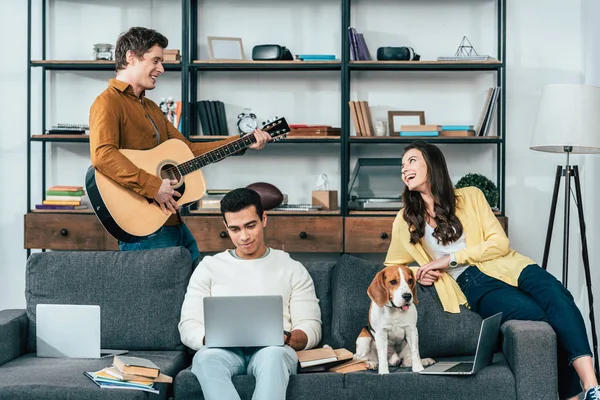 Három Multikulturális Hallgatók Kutyával Laptop Gitározni Nappaliban — Stock Fotó