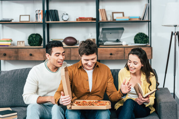 Трое смеющихся мультикультурных друзей сидят на диване с пиццей в гостиной
