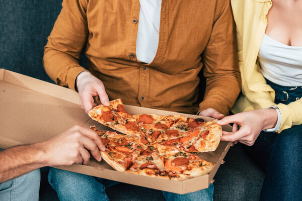 Cropped view of hree joyful friends sitting on sofa and eating pizza together