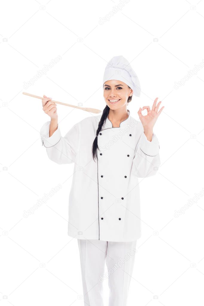 Smiling chef in uniform holding wooden spatula and showing okay sign isolated on white