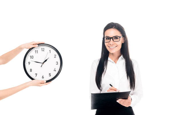 Businesswoman in glasses writing in clipboard near clock isolated on white