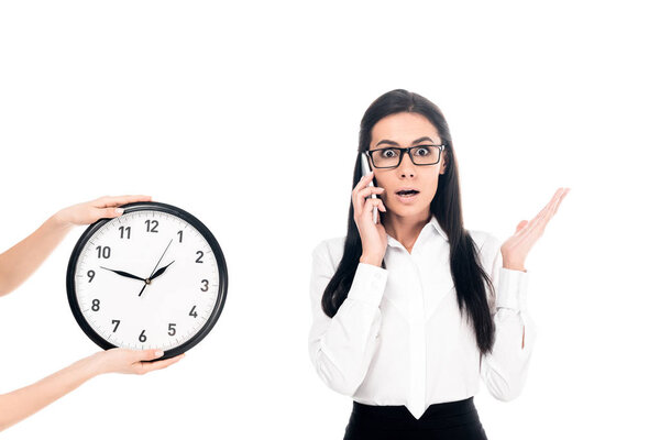 Shocked businesswoman talking on smartphone near clock isolated on white