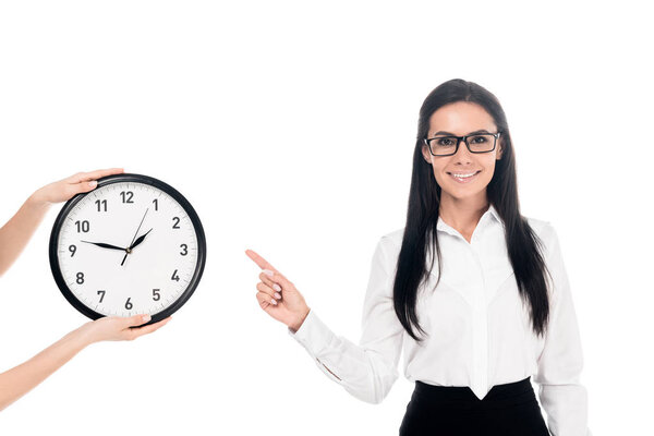 Smiling businesswoman in glasses pointing with finger at clock isolated on white