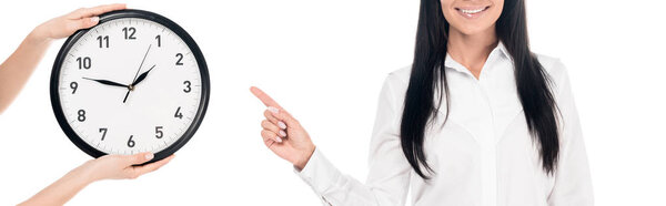 Panoramic shot of smiling businesswoman in glasses pointing with finger at clock isolated on white